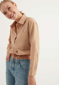 Camicia button-up beige chiaro con tasca, maniche lunghe e vita regolabile con cordoncino, abbinata a jeans azzurri. Tessuto liscio.