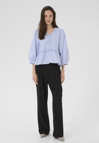 Bluza bleu cu dungi, cu decolteu în V cu volane, mâneci cu balon și talie cu peplum, asortată cu pantaloni negri cu talie înaltă și pantofi bej cu toc.