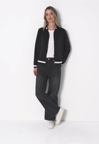 Czarna kurtka bomberka z białymi prążkowanymi akcentami, zestawiona z ciemnymi spodniami o wysokim stanie i białymi trampkami. Model stoi na jednolitym tle.