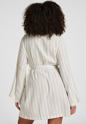 Donna con capelli ricci che indossa un abito crema fino al ginocchio, a maniche lunghe, con sottili righe verticali blu e una cintura in vita, vista di spalle.