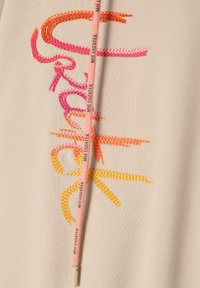 Tela beige con bordados multicolores en rosa, naranja y amarillo. El cordón presenta texto impreso y un acabado texturizado.