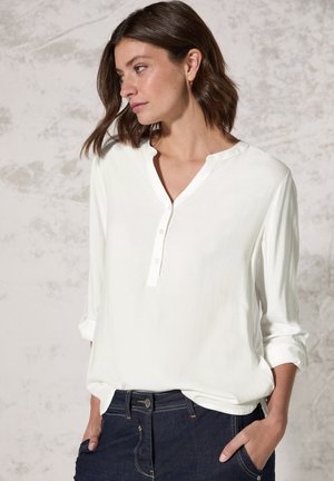 Lichte witte langemou-shirt met een v-hals en drie knopen bij de kraag. Gladde textuur, relaxte pasvorm, gestyled met donkere jeans.