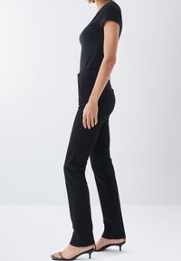 Femme portant un pantalon noir ajusté, un haut noir à manches courtes et des sandales noires à talons bas, debout devant un fond clair uni.