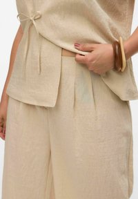 Haut en lin beige avec cordon de serrage et pantalon large plissé. Texture lisse, couleur neutre, complétée par un bracelet en bois au poignet.