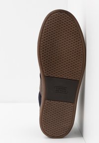 Hnědá guma podešve s texturovaným gripem; horní materiál v námořnické modři s koženým detailem a znakem. Profil podešve je plochý bez zvýšení.