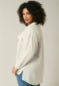 Light beige button-up shirt with a textured fabric, featuring a single chest pocket, long sleeves, and a curved hem. Paired with blue jeans.