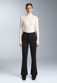 Hvid striktrøje med høj krave og mønstret tekstur; kombineret med sorte, udslåede bukser med sidelommer med lynlås og lukning med to knapper.