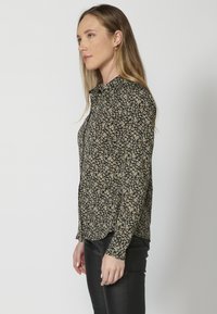Blusa nera con piccolo motivo floreale giallo, maniche lunghe, apertura frontale con bottoni e vestibilità aderente. Il tessuto appare liscio e leggero.