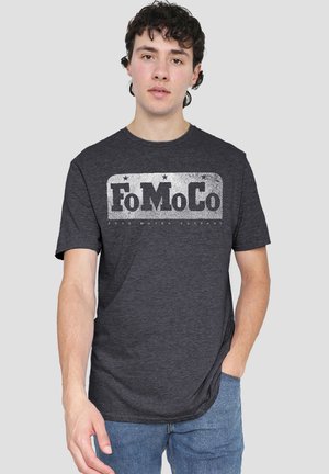 Junger Mann mit dunklen lockigen Haaren, der ein dunkelgraues FoMoCo-Logo-T-Shirt und blaue Jeans trägt und mit einer Hand in der Tasche steht.