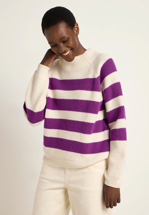Knit sweater featuring bold purple and white stripes, a round neckline, and ribbed cuffs, paired with light-colored pants.