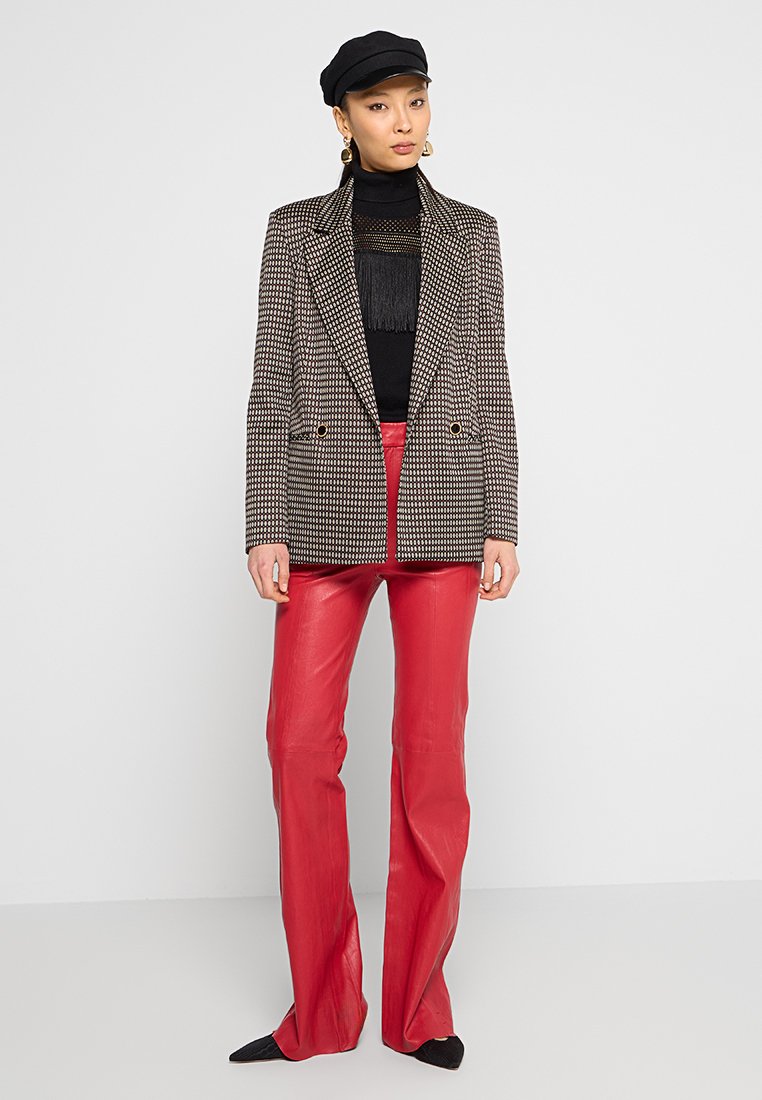 Eine Frau trägt eine schwarze Mütze, ein gemustertes Blazer, ein schwarzes fransiges Oberteil, rote Lederhosen und schwarze spitze Schuhe und steht vor einem weißen Hintergrund.