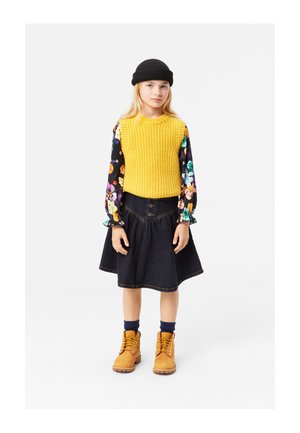 Geel gebreid trui, bloemen lange mouwen shirt, donker denim rok met knopen, en bruine veterschoenen met gele veters. Zwarte beanie.