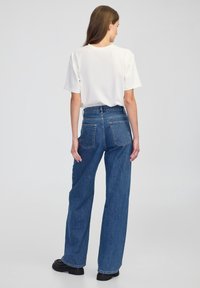 Overmaat wit T-shirt gecombineerd met hooghangende, wijde blauwe spijkerbroeken. Kenmerken zijn onder andere een klassiek ontwerp en achterzakken. Zwarte voetbedekking.