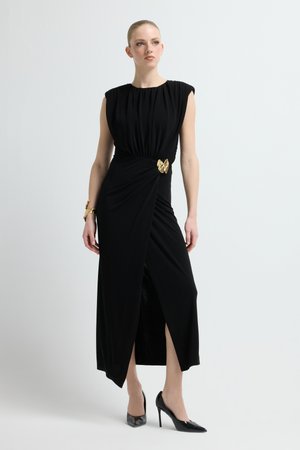 Czarna sukienka z drapowanym gorsetem, krótkimi rękawami i rozcięciem z przodu. Złota broszka w kształcie motyla podkreśla talię. Modelka ma na sobie czarne szpilki.