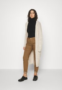 Cardigan longo bege com malha texturizada, usado sobre um suéter de gola alta preto, combinado com calças castanhas de corte justo e sapatos pretos de deslizar.