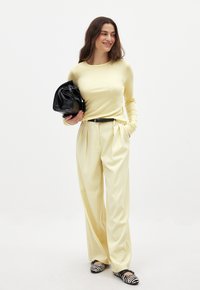 Helles gelbes Langarmoberteil, hochtaillierte gelbe Faltenhose, schwarzer Gürtel, schwarze Handtasche und schwarz-weiße gestreifte Slipper.