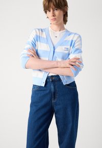 Light blue and white striped cardigan with a v-neck, button front, and logo patch, paired with high-waisted blue jeans.