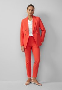 Korallenroter, doppelt gegenteiliger Blazer und passende, eng geschnittene Hosen, kombiniert mit einem weißen Top mit Schalkragen und weißen, schmalen Absatzsandalen. Strukturierter Stoff.