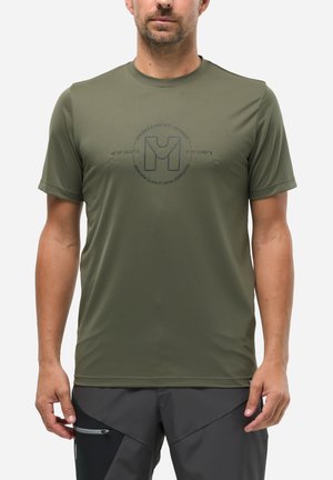 Homme portant un t-shirt kaki à manches courtes avec un logo centré sur la poitrine et un pantalon de plein air gris foncé avec une poche zippée.