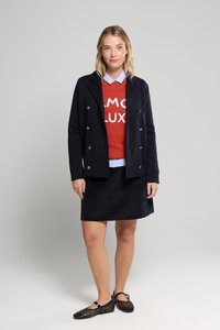 Veste marine à double boutonnage, pull rouge avec texte blanc, chemise col bleu clair, jupe noire et chaussures en maille noires.