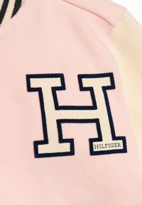 Rosa sweatshirt med kontrasterande beige ärmar, som har en stor broderad "H"-logotyp i kräm och marinblått, tillsammans med "HILFIGER"-etiketten.