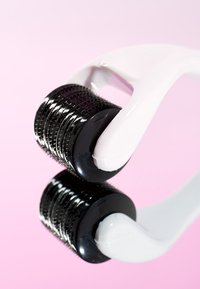 Derma roller med ett vitt plastgrepp och en svart roterande rulle täckt med fina nålar, reflekterande på en rosa bakgrund.