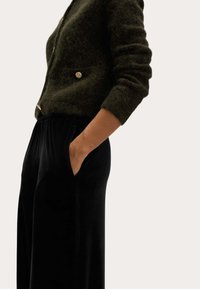 Ciemnozielony, teksturowany sweter z dzianiny z guzikami w złotym odcieniu, zestawiony z czarnymi, plisowanymi spodniami o szerokich nogawkach z bocznymi kieszeniami.