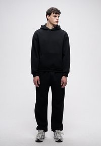 Felpa oversize nera in cotone con tasca frontale, abbinata a pantaloni della tuta neri coordinati. Il modello indossa sneakers grigie con accenti bianchi.