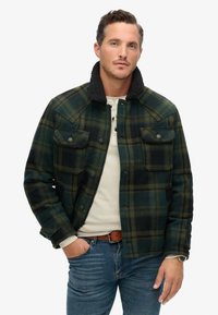 Grün- und schwarzkariertes Jackett mit Sherpa-Kragen, Knopfklappen und zwei Brusttaschen, getragen über einem hellen Hemd und blauen Jeans.