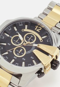 Orologio in metallo con quadrante nero, dettagli dorati e funzionalità cronografo. Bracciale in acciaio inossidabile e tono dorato. Grandi numeri e indicatori.