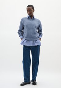 Siv krošnjati pulover preko modro-belih črtastih srajc s čipkastim robom, v kombinaciji s širokimi temno modrimi džins hlačami in črnimi loaferji.