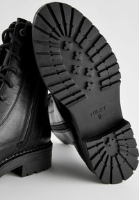 Svarta läderankelboots har en texturerad gummisula med detaljerat mönster. Snörningssystem och siddragkedja för enkel användning.