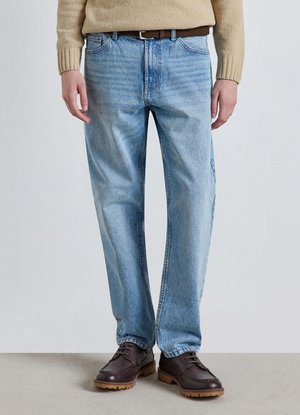 Persona che indossa jeans di denim celeste chiaro, cintura marrone, maglione beige e scarpe marrone scuro con lacci, in piedi su un pavimento grigio.