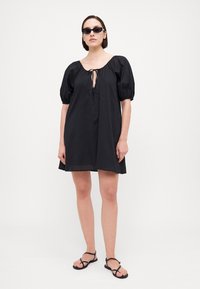 Μαύρο κοντό φόρεμα με χαλαρή εφαρμογή, φουσκωτά μανίκια και λεπτομέρεια με ζωνάκι στον λαιμό. Ελαφρύ ύφασμα με λεία υφή.