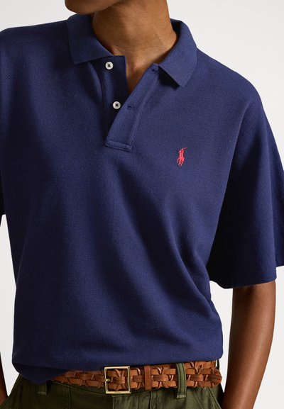 Μπλε ναυτικό πόλο πουκάμισο από υφασμένο ύφασμα, με τριπλό κούμπωμα με κουμπιά και μικρό κόκκινο λογότυπο στο αριστερό στήθος, φορεμένο με υφαντή καφέ ζώνη.