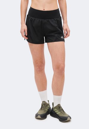 Γυναίκα που φοράει μαύρο αθλητικό σορτς, λευκές κάλτσες μέχρι τον αστράγαλο και πράσινα-μαύρα παπούτσια τρεξίματος, στέκεται μπροστά σε απλό φόντο.