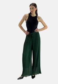 Zwarte mouwloze top gecombineerd met groene geplooide wijdere broeken met trekkoord in de taille en zwarte platte schoenen.