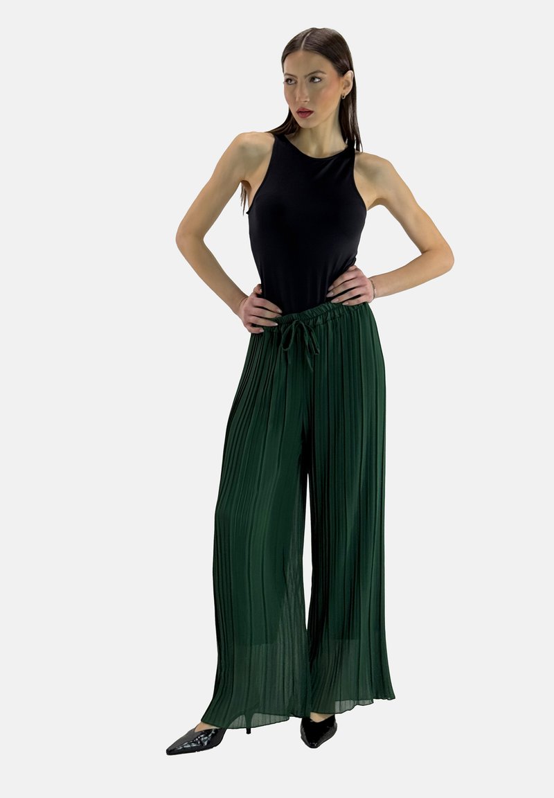 Zwarte mouwloze top gecombineerd met hooggesneden, geplooide groene broeken. De broeken hebben een trekkoord in de taille en een vloeiende drape.