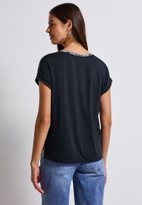 Schwarzes Kurzarmoberteil mit umgeschlagenen Ärmeln und einem gemusterten Rückensteg. Kombiniert mit hellblauen Jeans, von hinten gezeigt.
