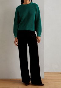 Zielony prążkowany sweter z dzianiny z długimi rękawami w połączeniu z czarnymi, szerokimi spodniami z aksamitu. Strój cechuje się luźnym krojem i miękką fakturą.