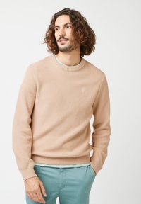 Beige strukturierter Pullover mit geripptem Ausschnitt und Bündchen. Verfügt über ein kleines Logo auf der Brust. Kombiniert mit hellblauen Chinos.