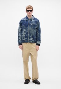 Chaqueta de denim con patrón de camuflaje azul y cierre de cremallera, combinada con pantalones de pierna ancha beige y zapatos negros sin cordones. La modelo lleva gafas de sol oscuras.