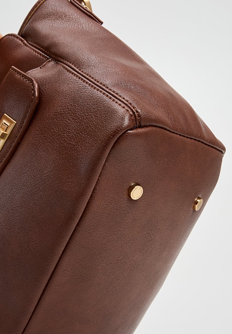 Sac à main en cuir marron avec une forme structurée, des accents en métal doré, des coutures visibles et des pieds en métal protecteurs à la base.