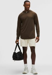 Hombre con camisa marrón con capucha y mangas largas y pantalones cortos color crema, sosteniendo una bolsa de deporte negra, de pie contra un fondo liso y claro.