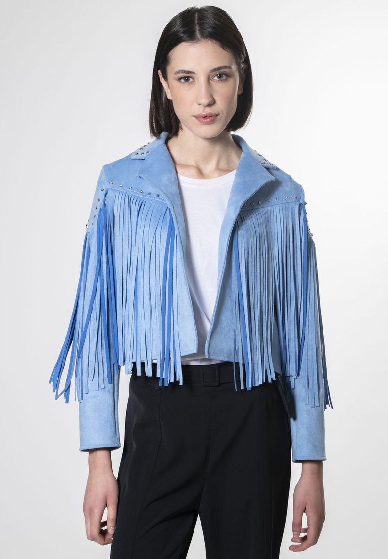 Giacca cropped in camoscio blu con dettagli di frange lunghe, borchie argentate sulle spalle, abbinata a una camicia bianca e pantaloni neri.