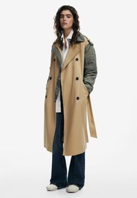 Beige trenchcoat met olijfgroene mouwen, dubbele knopen, grote knopen en een ceintuur in de taille, gedragen over een wit shirt en donkere jeans.