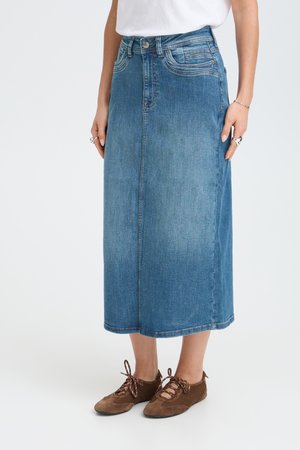 Kvinde iført en blå denim midi-nederdel kombineret med brune snørebåndssko og en hvid top, stående foran en ensfarvet baggrund.