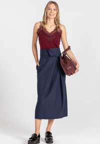 Burgundowa koszulka na cienkich ramiączkach z koronką, zestawiona z granatową spódnicą kopertową z guzikami. Modelka trzyma bordową zamszową torebkę i nosi czarne buty.