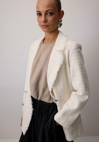 Biely tweed blazer s rozstrapkvanými hranami, svetlo béžová blúzka pod ním a čierna sukňa so zberaním. Odvážne strieborné gombíky ako akcenty na blazeri.