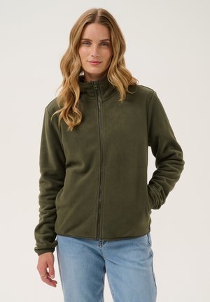 Jeune femme aux cheveux clairs et ondulés, portant une veste polaire zippée vert olive et un jean bleu clair, debout avec une main dans la poche.